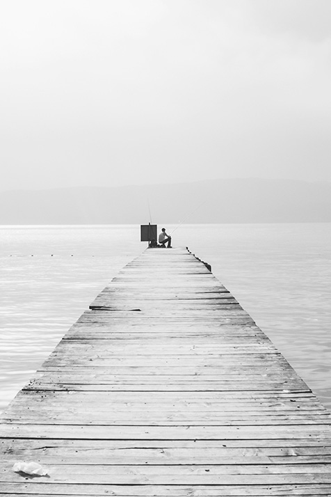 A pier stretches into the distant ocean and a single person sits at the end of it (black & white)