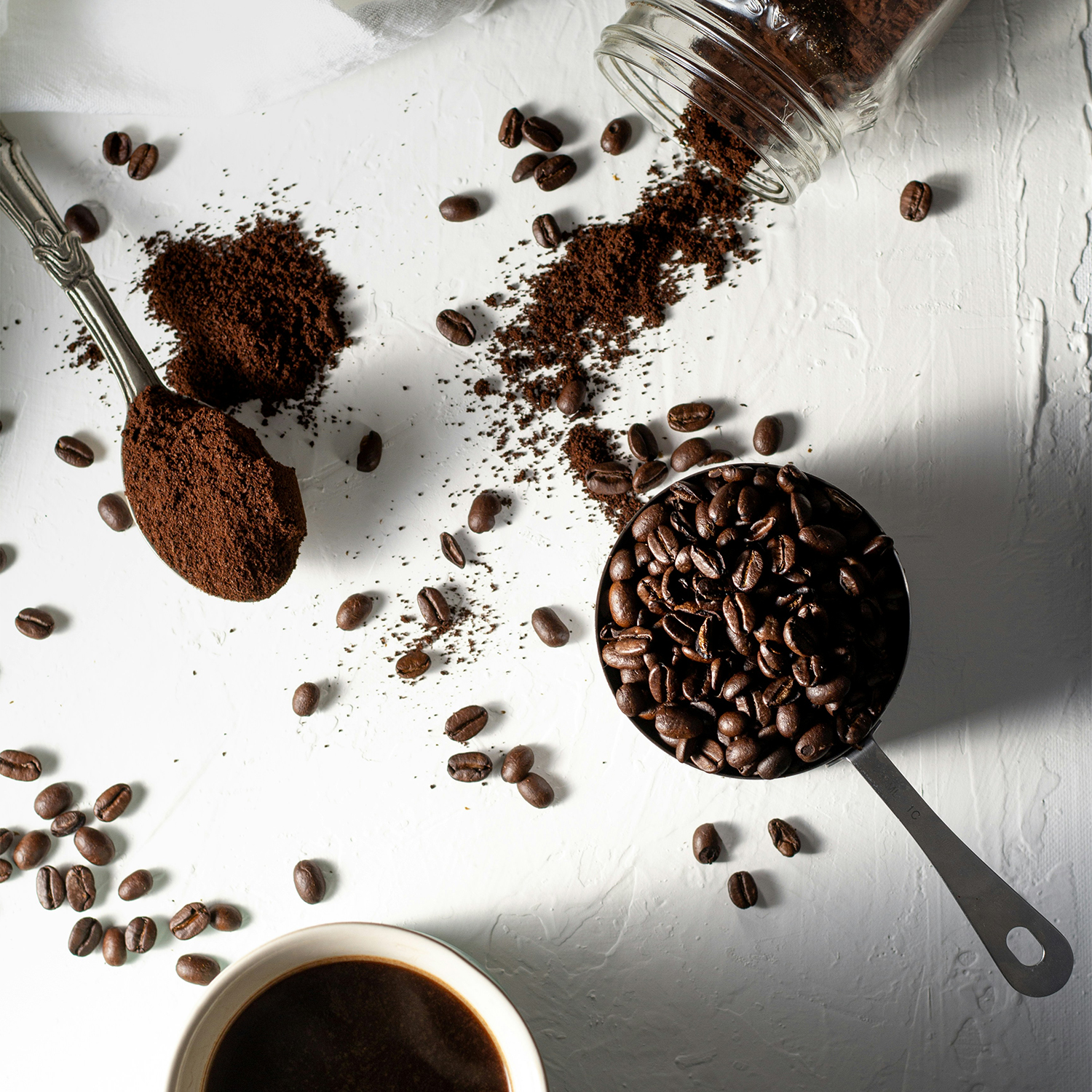 Measuring cup full of coffee beans next to a tipped over jar of coffee grounds next to a cup of cpffee
